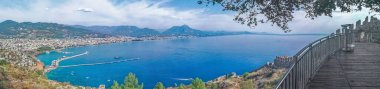 Türkiye 'nin Alanya Şatosu' ndaki manzaralı bir gözlem güvertesinden geniş bir panoramik manzara, Akdeniz ve Alanya kıyı şeridine bakıyor. Sahne, okyanusun güzelliğini, uzak dağları ve tarihi kale duvarlarını birleştirir.