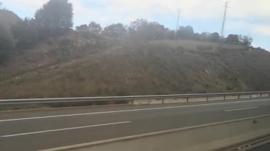 Otobüsün penceresinden bakıldığında, İspanya 'nın Katalonya kentindeki sonbahar manzaraları, kurumuş bitki örtüsü ve ekili tarlalarla kaplı tepeler görülüyor. Video 22 saniye. İki şeritli yol, parçalı bulutlu bir gökyüzünün altında metal bir güvenlik bariyeri içeriyor.