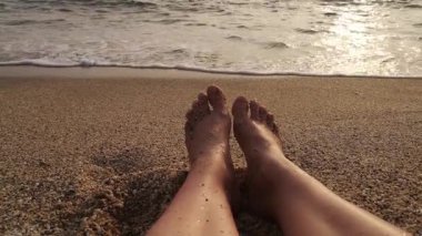 Dişi ayaklar kumsalda dinlenirken ayak parmakları havada su yavaşça kıyıda dolaşır. 4K video 28 saniye. Gün batımının altın rengi ıslak kumu yansıtır ve sakin bir sahil atmosferi yaratır.