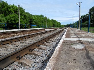 Güneşli bir günde, orman kuşağı arasında demiryolunun renkli bir fotoğrafı. Demiryolu rayları, yaya platformları, yeşil ağaçlar, güzel mavi bir gökyüzü ve ufukta iki adam yürüyüp gidiyor.
