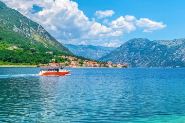 Karadağ 'ın Prcanj kıyı kenti yakınlarındaki Kotor Körfezi' nin sakin sularında gezinen bir tur teknesinin arka planında manzaralı dağlar ve manzaralı evler bulunuyor. Mükemmel bir yaz seyahati.