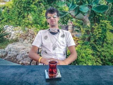 Bir adam sakin bir açık hava ortamında bir masada oturuyor, canlı yeşilliklerle çevrili bir fincan çayın tadını çıkarıyor. Rahatlama anı, Türkiye 'nin Alanya kentine yaptığı yolculuk sırasında barışçıl bir mola yakaladı.