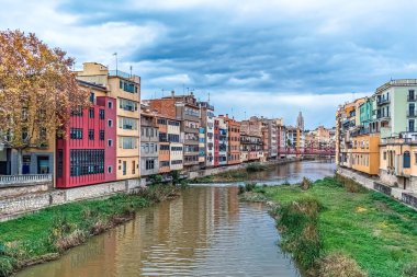 İspanya 'nın Girona kentinin panoramik manzarası, Onyar Nehri boyunca renkli evleri gösteriyor. Sonbahar ağaçları ve bulutlu gökyüzü bu pitoresk şehrin cazibesini artırıyor.