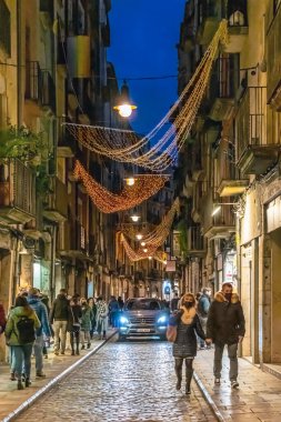 Girona 'daki Carrer de les Ballesteries' in gece manzarası ve Noel süslemeleri. Canlı ortaçağ caddesi yayalarla, aydınlanmış dükkanlarla dolu. Altın ışık çelenkleri büyülü bir ortam yaratır. Girona, İspanya - 27 Kasım 2021