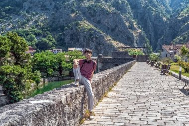 Karadağ 'ın manzaralı Kotor tahkimatları boyunca taş bir duvarda dinlenen bir adam. Arka planda engebeli dağlar ve yeşillikler, barışçıl bir atmosfer ve tarihi mimariyle sergileniyordu.