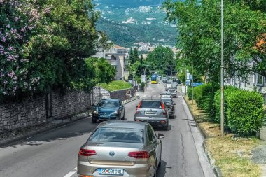 Budva 'da ağaçlarla kaplı, yemyeşil bir cadde boyunca giden çok sayıda arabalı manzaralı bir yol. Dağlar ve konut binaları uzaktan görülebiliyor ve manzarayı aydınlatıyor. Budva, Karadağ - 18 Haziran 2021