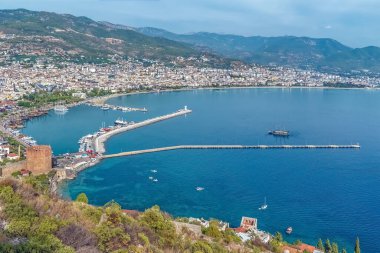 Alanya limanı ve Kızıl Kule 'nin panoramik manzarası. Şehrin ufuk çizgisi ve arka planında dağlar var. Huzurlu Akdeniz suları marina ve tarihi mimariyi ön plana çıkarıyor.