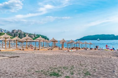 Karadağ 'ın Budva kentindeki Plaza Zuplica plajında güneşli yataklar ve şemsiyelerle kumlu sahil boyunca dizilmiş rahatlatıcı bir plaj sahnesi. Arka planda Sveti Nikola Adası, sakin Adriyatik Denizi 'nde yelken açan bir yatla görülebiliyor. Budva, Karadağ - 18 Haziran, 2