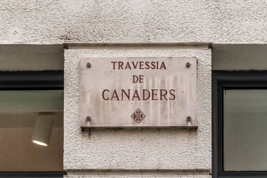 Eski Girona kasabasında beyaz dokulu duvara asılmış kırmızı harflerle yazılmış, Travessia de Canaders yazılı Stone Caddesi plakası. Tarihi şehir merkezinde dekoratif amblemli geleneksel Katalan sokak tabelası. Girona, İspanya - 27 Kasım 2021