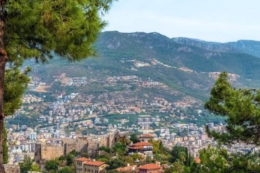 Alanya Kalesi 'nden Alanya şehrinin manzarası, çevredeki dağları ve şehri gösteriyor. I Love Alanya tabelası yamaçta görünüyor, bölgenin panoramik güzelliğini vurguluyor.