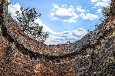 Türkiye 'nin Alanya Şatosu' ndaki bir kuleden mavi bulutlu gökyüzüne ve bir ağaca açılan oval bir delikten yukarıya bakan bir manzara. Antik taş duvarlar yukarıdaki doğal manzarayı çerçeveler.