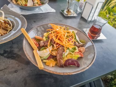Türkiye 'nin Alanya kentindeki açık bir masada taze sebze salatası ve bir bardak çay ile servis ediliyor. Tabakta canlı malzemeler yer alıyor, manzaralı ve huzurlu bir yemek deneyimi sunuyor.