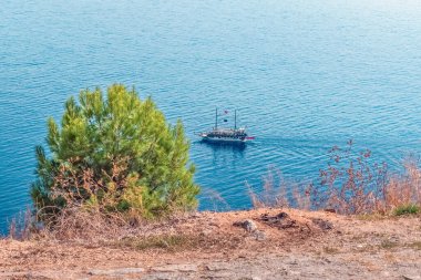 Türkiye 'nin Alanya kenti yakınlarındaki Akdeniz' in mavi sularında huzur içinde yüzen bir tekne görülüyor. Ön plandaki yemyeşil çam ağacı sakin okyanusla çelişiyor ve sakin bir manzara yaratıyor.