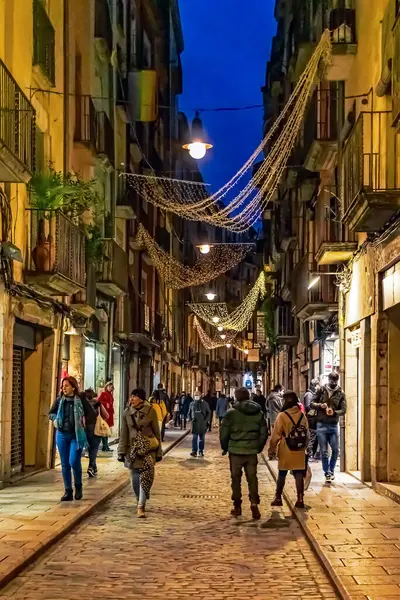 Ortaçağ Carrer de les Ballesteries caddesi Girona 'da Noel ışıklarıyla süslenmiş. İnsanlar akşam saatlerinde tarihi binalar arasında kaldırımda yürüyorlar. Girona, İspanya - 27 Kasım 2021