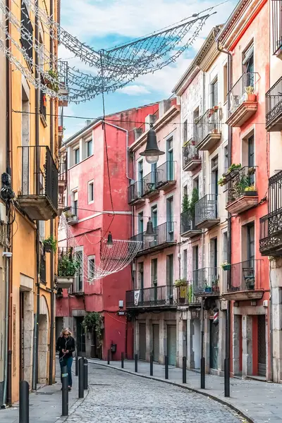 Girona 'da demir balkonları ve saksı bitkilerinin yer aldığı pembe, mercan ve sarı binaları olan renkli dar bir cadde. Noel süsleri başımızın üstünde asılı. Çakıl taşı yolu. Girona, İspanya - 28 Kasım 2021