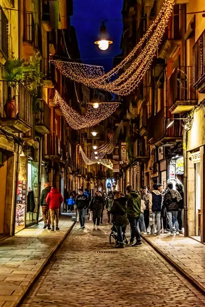 Girona 'daki tarihi Carrer de les Ballesteries caddesini aydınlattı. Noel ışıkları başınızın üstünde kıvrımlı desenler oluşturdu. İnsanlar geceleri kaldırım taşı caddesinde alışveriş yapıp yürüyorlar. Girona, İspanya - 27 Kasım 2021