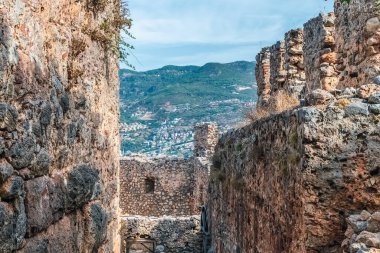 Alanya Kalesi 'nin yıpranmış taş duvarları Türkiye' nin dağlık arazisinin uzak bir manzarasını çerçeveliyor. Antik kale, çatlakların arasından çıkan kaya ve bitkilerle çürüme belirtileri gösteriyor.