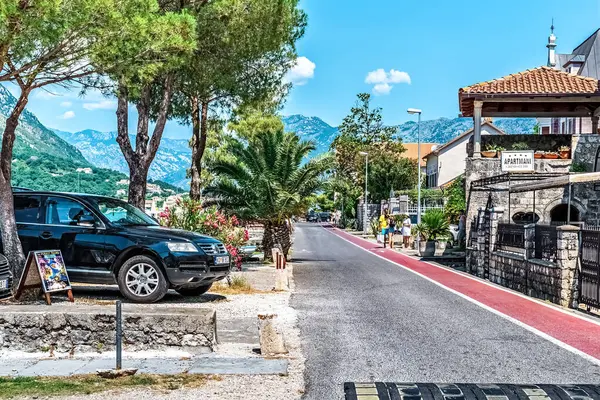 Kotor 'da güneşli bir günde park halindeki arabaların, palmiye ağaçlarının ve yayaların yürüdüğü büyüleyici bir cadde. Arkaplan manzaralı dağları da içeriyor, bu da onu huzurlu bir yaz manzarası yapıyor. Kotor, Karadağ - 18 Haziran 2021