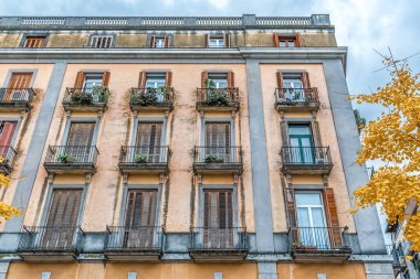 PI 'da çok katlı bina cephesi. De la Independencia, 18, Girona, İspanya. Binanın içinde balkonlar, ahşap kepenkler, saksılar ve sonbahar yapraklı görünür yaşlanma detayları yer alıyor.