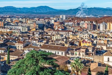 Arka planda yoğun şehir mimarisi, terasotta çatıları ve Pirene dağları olan Girona şehrinin panoramik hava manzarası. İspanya 'nın Katalonya şehrinde açık bir kış gününde sanayi dumanı yükseliyor