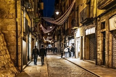 Girona 'daki Carrer de les Ballesteries' in akşam manzarası ve Noel ışıkları süsü. İnsanlar ortaçağ dar sokaklarında tarihi taş binalar ve aydınlatılmış vitrinlerle yürüyorlar. Girona, İspanya - 27 Kasım 2021