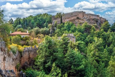 Türkiye 'nin parçalı bulutlu gökyüzünün altında yemyeşil ve antik taş duvarlarla çevrili Alanya Kalesi' nin pitoresk bir manzarası. Tarihi tahkimatlar doğal manzarayla güzel bir şekilde harmanlanmış.