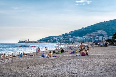 Budva 'daki Plaza Zuplica plajında insanların güneşlendiği ve Adriyatik Denizi' nin tadını çıkardığı canlı bir yaz sahnesi. Arka plandaki kıyı kasabası ve dağlar manzaraya eklenir. Budva, Karadağ - 18 Haziran 2021