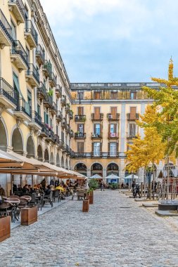 Geleneksel neoklasik mimarisi ve açık kafe terasları ile Girona 'daki tarihi Placa de la Independencia. Sonbahar sahnesi, kaldırım taşı meydanı, sarı yapraklar, ve tarihi binaların kemerli sütunları. Girona, İspanya - 27 Kasım 2021