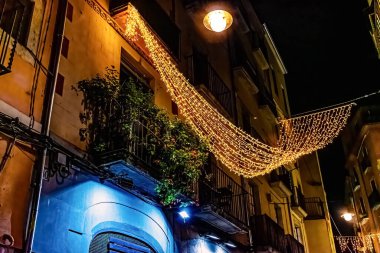 İspanya 'nın tarihi Girona kentindeki atmosferik gece sahnesi. Geleneksel cadde sıcak sarı Noel ışıklarıyla süslenmiş. Mavi ışıklı giriş ve altın peri ışıkları arasındaki zıtlık ortaçağ Katalan bölgesinde büyülü bir festival ortamı yaratır.