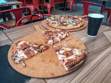 Sıradan bir pizzacının ahşap tahtalarında servis edilen iki lezzetli pizza ve arka planda parlak kırmızı metal sandalyeler. Dilimlenmiş bir pizza, ikinci tam pizza ise renkli sebze ve et karışımı ile kaplı.