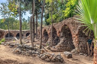 Türkiye 'nin Alanya Şatosu' nda ağaçlar ve yeşilliklerle çevrili antik taş ve tuğla kemerler. Yapıda tarihi mimari ve doğal güzellikler sergilenirken, ön planda kazı çalışmaları görülebiliyor.
