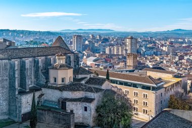 Girona şehrinin panoramik manzarası. İspanya 'nın Girona şehrinde bulunan Sant Domenec Manastırı. Terracotta çatıları olan tarihi taş binalar, berrak mavi gökyüzünün altındaki uzak dağlara uzanan modern şehir manzarasıyla tezat oluşturuyor.