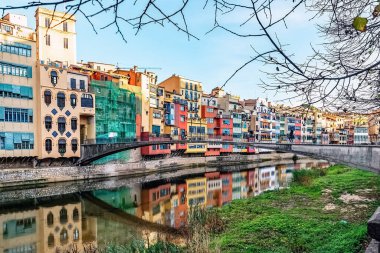 İspanya 'nın Girona kentindeki Pont d en Gomez' deki sularda yansıyan Onyar Nehri boyunca renkli evler. Bu ikonik nehir kenarındaki binaların canlı cephelerinde çıplak kış dalları tarafından çerçevelenmiş geleneksel Katalan mimarisi sergileniyor.