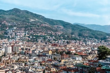 Türkiye 'nin Alanya kentinin panoramik manzarası, kentin binalarının manzaraya yayılması ve tepenin üzerindeki Alanya tabelasının görünmesi. Arkaplanda açık bir gökyüzünün altındaki yeşil dağlar var.