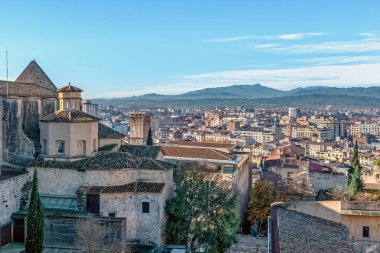Sant Domenec Manastırı 'ndan Girona şehrinin panoramik manzarası, İspanya' nın Pirenes dağlarının arka planına karşı tarihi mimari sergileniyor. Süpürme şehri, açık mavi bir gökyüzü altında antik yapılar ve modern binaların bir karışımını ortaya çıkarıyor.