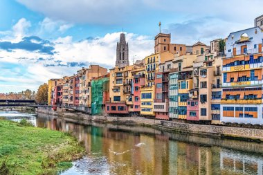 İspanya 'nın Girona kentindeki Onyar Nehri boyunca uzanan renkli binalar, nehir manzaralı ve parçalı bulutlu bir gökyüzü altında tarihi mimariye sahip. Sudaki yansımalar resim gibi bir sahne yaratır.