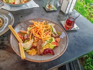 Türkiye 'nin Alanya kentindeki bir masada, renkli bir sebze salatası ve geleneksel Türk çayı servis ediliyor. Taze yemek, huzurlu bir bahçe ortamında, rahatlatıcı bir öğle yemeği için mükemmeldir.