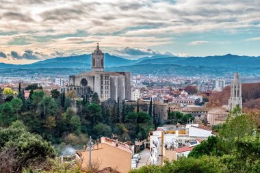 İspanya, Girona 'nın panoramik manzarası, Gotik Girona Katedrali' nin eski kasabanın üzerinde yükselmesi, sonbahar yeşillikleri ve modern şehir, Pirene dağlarına ve dramatik bulutlu gökyüzüne karşı kurulmuş.