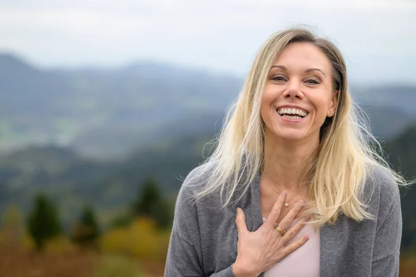 Portrait of an extremely happy and laughing middle aged blond woman looking straight into the camera on top of a mountain and cloudy sky
