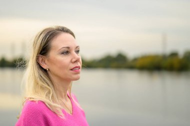 Pembe süveter ve pembe ruj süren orta yaşlı muhteşem bir kadın, sonbaharda bir nehrin ya da gölün önünde duruyor.