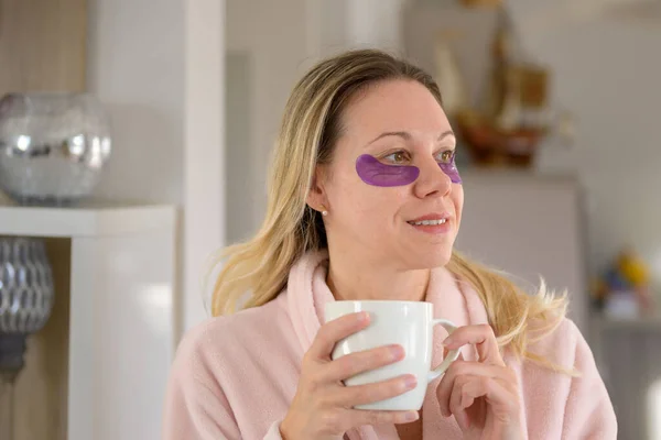 Side view of an smiling attractive middle aged woman in her pink bathrobe with eye patches holding a cup of coffee in the living room