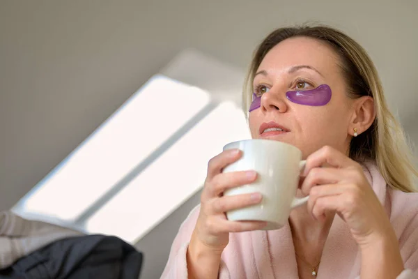Side view of an attractive middle aged woman in her pink bathrobe with eye patches holding a cup of coffee in front of a window