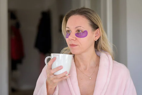Side view of an attractive middle aged woman in her pink bathrobe with eye patches holding a cup of coffee