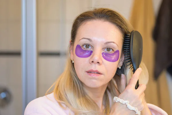 Morning routine of an attractive middle-aged woman in her bathroom wearing eye pads combs her hair with a brush