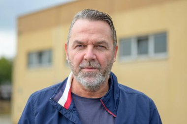 Orta yaşlı bir adam, sarı bir binanın önünde sert bir bakışla kameraya bakıyor.