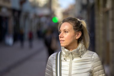 Hafif gülümseyen orta yaşlı, sarışın ve at kuyruklu çekici bir kadının yan görünüşü metalik bir ceket giyiyor ve omzunun üzerinden bir el çantası alışveriş caddesinde yürüyor.