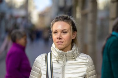 Nazikçe gülümseyen orta yaşlı, sarı saçlı, at kuyruklu çekici bir kadın metal renkli bir ceket giyiyor ve omzunda bir el çantası alışveriş caddesinde yürüyor.
