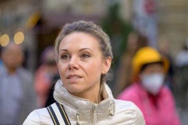 Neşeyle gülen orta yaşlı, sarı saçlı, at kuyruklu çekici bir kadın. Metalik bir ceket giyiyor. Omzunda bir çanta alışveriş caddesinde yürüyor.