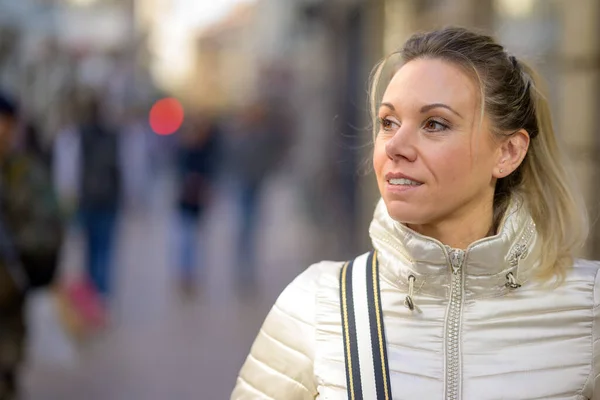 stock image Portrait of a gently smiling middle-aged attractive woman with blond hair and a ponytail wears a metallic-colored jacket and a handbag over her shoulder walks through a shopping street