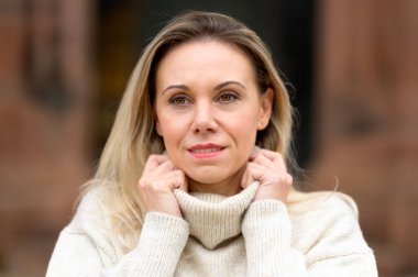 Çekici sarışın orta yaşlı bir kadın gülümsüyor ve elleri boğazlı eski kahverengi bir kiliseye bakıyor.